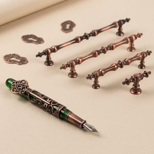 May include: A collection of antique-style hardware, including bronze-coloured handles, keyhole covers, and a decorative fountain pen with a green accent. The pen features intricate detailing, and the hardware has a vintage aesthetic.