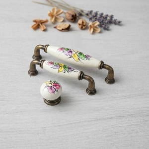 May include: Set of two white ceramic cabinet handles and a matching knob. The handles have a floral design in purple, yellow, and green. The hardware has a bronze finish. The knob is round with a similar floral pattern.