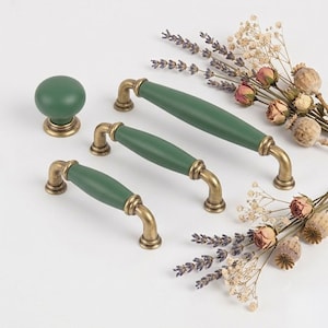 Pode incluir: Um conjunto de três puxadores de armário verdes e um botão de armário redondo a condizer. Cada peça apresenta um acabamento verde mate e liso no corpo do puxador ou botão, acentuado por bases de metal em tom de latão antigo nas junções.