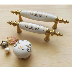 May include: Two white and gold cabinet pulls with a decorative floral design. The pulls have a gold base and ends. A matching white and gold cabinet knob is also present, along with a pearl earring and dried flower petals.