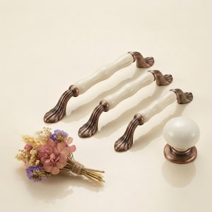 May include: A set of four cabinet pulls and a knob. The pulls have a white ceramic centre and bronze-coloured decorative ends. A small bouquet of dried flowers is also present. The hardware is designed for furniture.