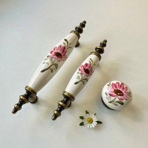 May include: Set of white ceramic cabinet pulls and a knob, each adorned with a pink floral design. The hardware features bronze-colored accents and a decorative shape. A small daisy is also present.