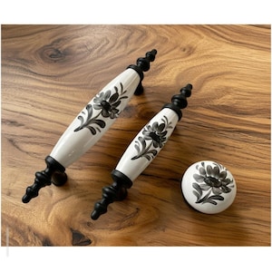 Könnte beinhalten: Zwei weiße und schwarze Schrankgriffe mit floralem Design und ein passender Knauf. Die Griffe sind länglich mit schwarzem Sockel und Zierknauf. Der Knauf ist rund. Das florale Design ist in Schwarz und Grau gehalten.