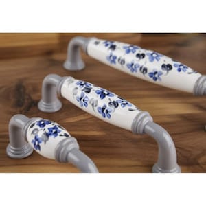Puede incluir: Tiradores de armario con diseño floral. Los tiradores tienen un centro de cerámica blanca con flores azules y soportes de metal gris. Están dispuestos sobre una superficie de madera.