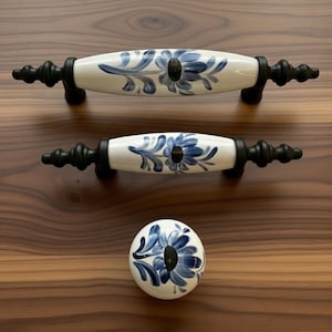 Może przedstawiać: Dwa podłużne ceramiczne uchwyty do szuflad oraz jedna okrągła ceramiczna gałka, wszystkie z białym tłem, ręcznie malowanymi niebieskimi wzorami kwiatowymi i czarnymi metalowymi okuciami. Uchwyty posiadają ozdobne zakończenia po obu stronach.