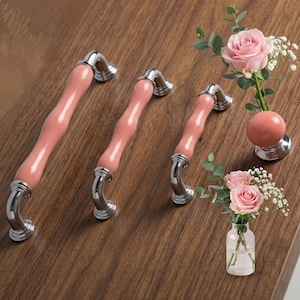 Op de afbeelding: Drie kastgrepen met een golvend, lichtroze middenstuk en zilverkleurige uiteinden worden getoond op een houten oppervlak. Een kleine vaas met roze rozen en groen is ook aanwezig en voegt een decoratief tintje toe.