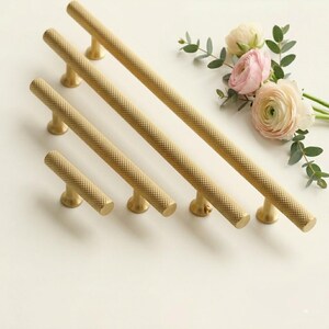 May include: A collection of gold-colored cabinet pulls with a textured diamond pattern. The pulls are arranged in a row, varying in length, and are mounted on a white surface. A bouquet of pink and white flowers with green leaves is in the background.