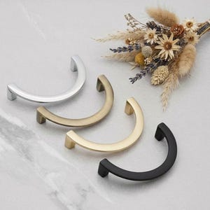 May include: Four curved cabinet pulls in silver, brass, gold, and black. The pulls are arranged on a marble surface, with a small bouquet of dried flowers in the background. These are ideal for kitchen or bathroom cabinets.
