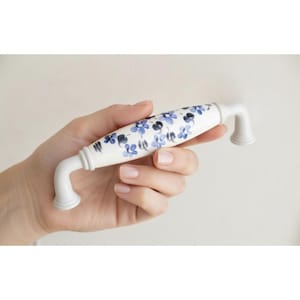 May include: White ceramic cabinet handle featuring a blue and dark blue floral pattern. The handle has a curved design with decorative bases. A hand is holding the cabinet handle.