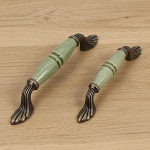 Può includere: Due maniglie per mobili in stile antico. Ogni maniglia presenta un centro in ceramica verde chiaro con una glassa craquelé, accentuato da due bande verdi più scure. Le maniglie hanno una base color bronzo e finiture decorative.