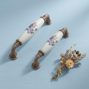 May include: Two white ceramic cabinet handles with bronze-coloured metal fittings. Each handle has a floral design in purple and green. A small bouquet of dried flowers rests on a light blue surface.