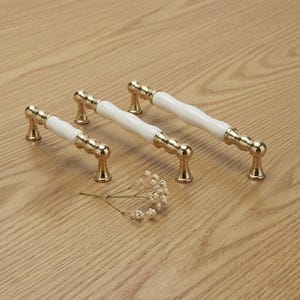 Könnte beinhalten: Drei Schrankgriffe mit weißer Mitte und goldfarbenen Akzenten. Die Griffe sind auf einer hellen Holzoberfläche angeordnet. Die Hardware hat ein klassisches Design, geeignet für Küchen- oder Badezimmerschränke.