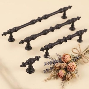 May include: A collection of black metal cabinet pulls in various sizes. The pulls have an ornate, vintage design with decorative detailing. A small bouquet of dried flowers and lavender is also present.