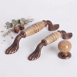May include: Two cabinet handles and a knob. The handles have a bronze-colored base with a decorative, curved design. The center section is a cream color with a crackle pattern. The knob is round with a matching cream and bronze color scheme.
