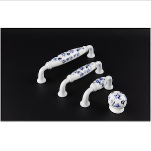 Puede incluir: Juego de herrajes de armario de cerámica blanca con estampado floral azul. Incluye dos tiradores y un pomo. Los tiradores tienen un diseño curvo y el pomo es redondo. El conjunto está sobre un fondo negro.