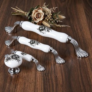 Può includere: Un set di maniglie per mobili e un pomello in ceramica bianca con un motivo floreale nero e grigio. Le maniglie hanno una base e un'impugnatura color argento. Il pomello è rotondo con un motivo floreale abbinato. Fiori secchi decorativi sullo sfondo.