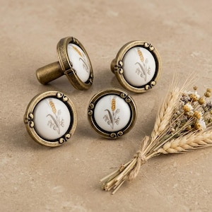 May include: Four antique brass cabinet knobs with white ceramic faces. Each knob features a wheat stalk design in yellow and brown. The knobs are arranged on a neutral surface, with a small bundle of wheat stalks in the foreground.
