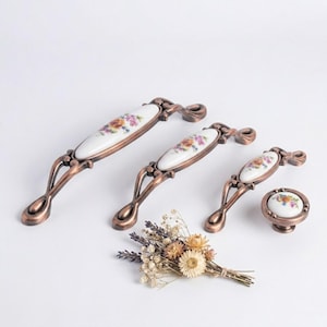 May include: A set of antique-style cabinet hardware featuring bronze-colored pulls and a knob. Each piece has a white ceramic center with a floral design in shades of pink, yellow, and orange. The hardware is arranged on a white surface.