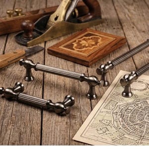 May include: Three dark metallic cabinet pulls with a fluted cylindrical center and rounded ends. The hardware features a polished finish and is arranged alongside a wooden block with an inlaid star pattern and a vintage-style map on paper.