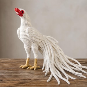May include: A handmade crochet bird sculpture in white with a red beak and eye details. The bird has long, flowing tail feathers and yellow feet, standing on a wooden surface. The sculpture is a unique decorative item.