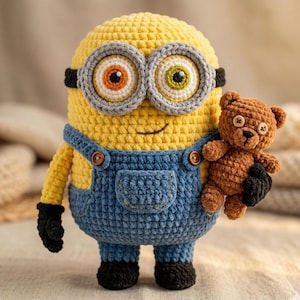 Könnte beinhalten: Eine gehäkelte Minion-Figur in Gelb mit blauen Latzhosen, schwarzen Handschuhen und Schuhen, die einen kleinen braunen Teddybären hält. Der Minion hat eine graue Brille und zwei Augen mit orangefarbenen und grünen Iris.