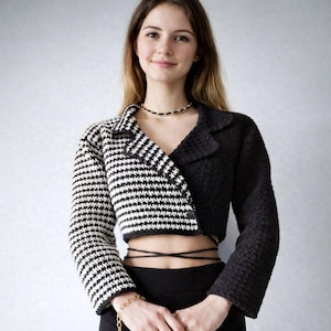 May include: A cropped blazer with a unique design. One side features black and white horizontal stripes, while the other is solid black. The blazer has a notched collar and long sleeves. The model is wearing a black skirt and a gold chain bracelet.