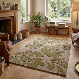 May include: A rectangular area rug with a floral design in shades of green and beige. The rug is placed on a wooden floor in a living room setting, with a fireplace, windows, and furniture visible in the background.