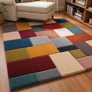 May include: A colorful patchwork rug with a geometric design. The rug features a variety of rectangular blocks in shades of blue, red, yellow, green, and neutral tones. The rug is placed on a hardwood floor, with a beige chaise lounge and bookshelf in the background.