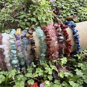 Puede incluir: Una colección de pulseras de virutas de piedras preciosas en varios colores, incluyendo morado, azul, verde y rojo. Las pulseras se exhiben sobre un cilindro cubierto de arpillera, rodeadas de follaje verde.