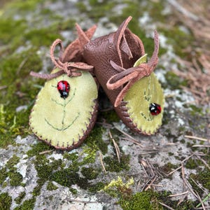 May include: Handmade baby moccasins. The moccasins are made of brown and light green leather. The light green leather is leaf-shaped with a stitched leaf design and a ladybug. The moccasins are tied with leather laces.