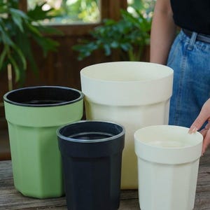 May include: A set of four cylindrical plant pots with a faceted exterior design. The collection includes one large cream pot, one medium green pot with a black interior, one medium black pot, and one small cream pot, all arranged on a wooden surface.
