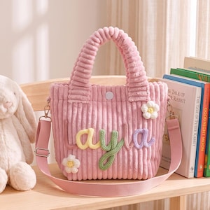 May include: A pink corduroy tote bag featuring the name Layla in colorful yarn lettering. The bag has two top handles, a detachable shoulder strap, a snap button closure, and two small white flower embellishments on the front surface.