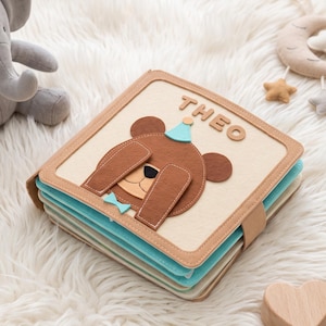May include: A square fabric book with a beige cover featuring a brown bear with a blue party hat and bowtie. The name THEO is stitched in brown letters above the bear. The book has layered pages in shades of blue and tan, secured with a tan strap.