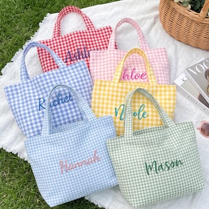 Op de afbeelding: Zes kleine draagtassen in verschillende gingham-kleuren, waaronder rood, roze, blauw, geel, lichtblauw en groen. Elke tas is aan de voorzijde voorzien van een gepersonaliseerde naam in cursief schrift. De tassen liggen op een witte stoffen ondergrond.