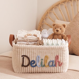 Puede incluir: Una cesta de almacenamiento rectangular de pana beige con asas de piel sintética marrón. El nombre Delilah está bordado en la parte delantera con letras de hilo multicolor. La cesta contiene ropa de bebé doblada, un mordedor y pañales.