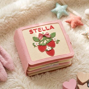Op de afbeelding: Een vierkant stoffen boek met een crèmekleurige kaft met de naam STELLA in rode letters boven een aardbeienapplicatie met groene bladeren, witte bloemen en een rood-wit geblokte strik, afgewerkt met een roze rand en gelaagde pagina's.