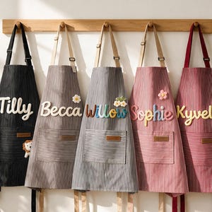 Könnte beinhalten: Fünf gestreifte Schürzen mit verstellbaren Nackenbändern und Vordertaschen, jede mit einem personalisierten Namen in erhabenen, bunten Buchstaben. Die Schürzen sind in Schwarz, Hellgrau, Dunkelgrau, Rosa und Rot an einer Holzwandleiste aufgehängt.