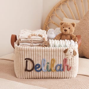 May include: A rectangular beige corduroy storage basket with brown faux leather handles. The name Delilah is embroidered in colorful yarn on the front. The basket is filled with folded baby clothes, a teddy bear, baby bottles, and diapers.