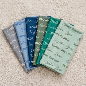 K&ouml;nnte beinhalten: Eine Sammlung von sechs gefalteten Stoffmustern in den Farben Grau, Blau, Marineblau, Olivgr&uuml;n, Smaragdgr&uuml;n und Salbeigr&uuml;n. Jeder Stoff zeigt ein sich wiederholendes Muster aus Namen, die in einer Mischung aus Blockschrift und Schreibschrift gedruckt sind.
