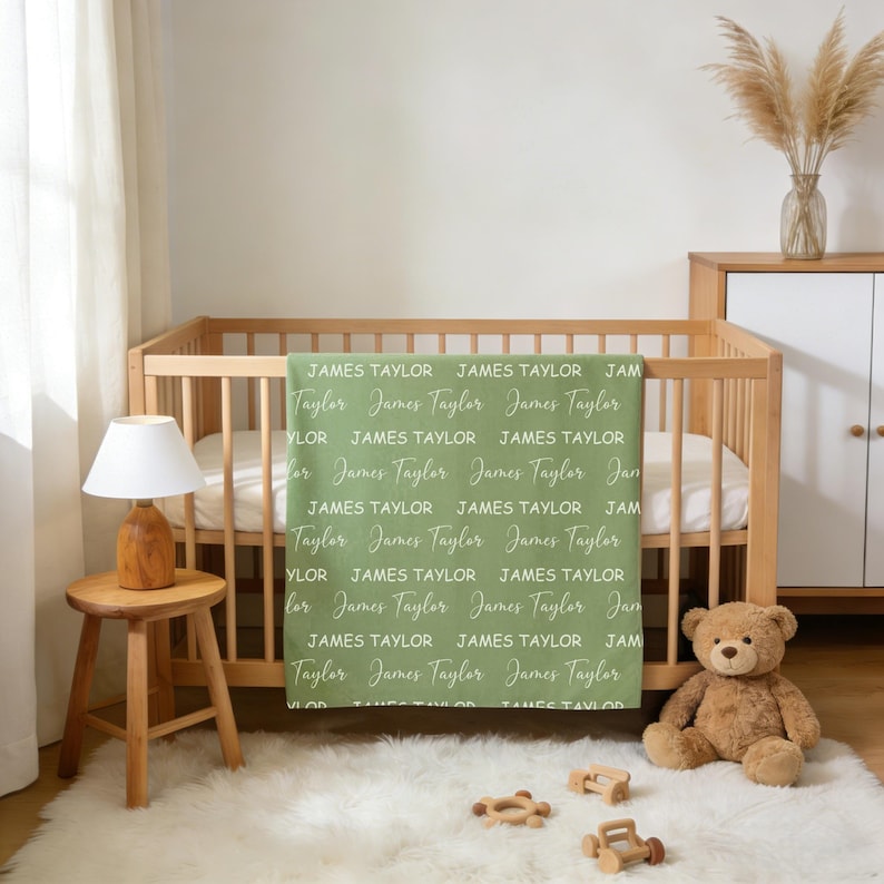K&ouml;nnte beinhalten: Eine salbeigr&uuml;ne Babydecke mit einem sich wiederholenden Muster des Namens James Taylor, gedruckt in abwechselnder Block- und Schreibschrift in Wei&szlig;. Die Decke h&auml;ngt &uuml;ber der Seite eines h&ouml;lzernen Kinderbetts in einem Kinderzimmer.