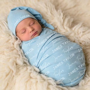 K&ouml;nnte beinhalten: Ein hellblaues Baby-Pucktuch-Set mit passender Knotenm&uuml;tze. Der Stoff ist mit dem Namen James Taylor in wei&szlig;er Schrift in Block- und Schreibschrift bedruckt. Das Pucktuch ist um einen schlafenden S&auml;ugling gewickelt.