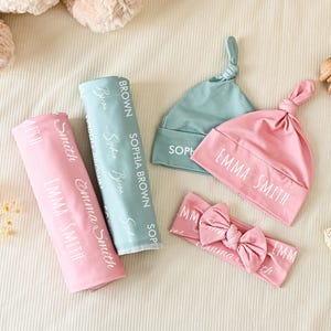 K&ouml;nnte beinhalten: Ein Set aus Babyzubeh&ouml;r mit einer rosa Wickeldecke und passender Knotenm&uuml;tze mit dem in Wei&szlig; gedruckten Namen Emma Smith, einer salbeigr&uuml;nen Wickeldecke und passender M&uuml;tze mit dem Namen Sophia Brown sowie einem rosa Stirnband mit Schleife.