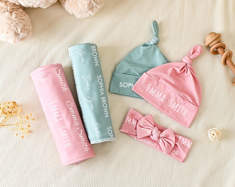 K&ouml;nnte beinhalten: Ein Set aus Babyzubeh&ouml;r mit einer rosa Wickeldecke und passender Knotenm&uuml;tze mit dem in Wei&szlig; gedruckten Namen Emma Smith, einer salbeigr&uuml;nen Wickeldecke und passender M&uuml;tze mit dem Namen Sophia Brown sowie einem rosa Stirnband mit Schleife.