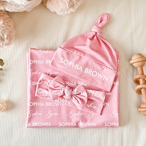 K&ouml;nnte beinhalten: Eine rosa Baby-Wickeldecke, eine Knotenm&uuml;tze und ein Schleifen-Stirnband, alle mit dem wei&szlig;en Text "SOPHIA BROWN" in verschiedenen Schriftarten bedruckt. Das Set ist zusammen abgebildet und zeigt den passenden Stoff und das personalisierte Design.