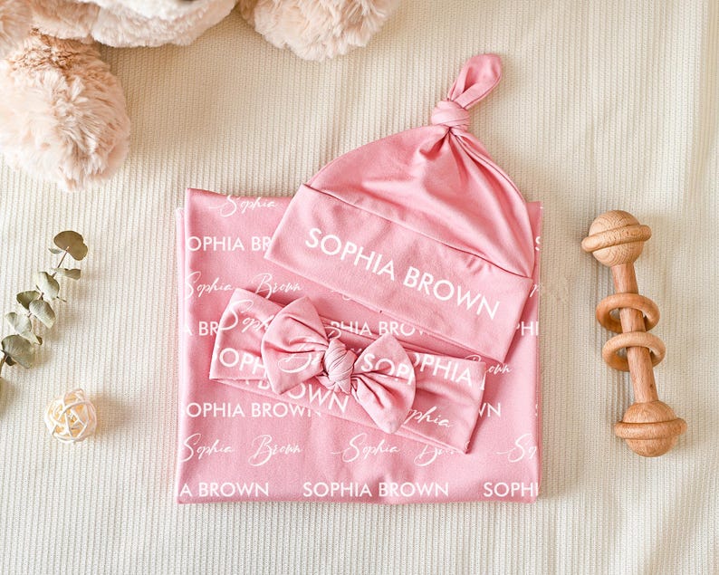 K&ouml;nnte beinhalten: Eine rosa Baby-Wickeldecke, eine Knotenm&uuml;tze und ein Schleifen-Stirnband, alle mit dem wei&szlig;en Text "SOPHIA BROWN" in verschiedenen Schriftarten bedruckt. Das Set ist zusammen abgebildet und zeigt den passenden Stoff und das personalisierte Design.
