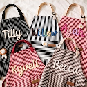 Könnte beinhalten: Fünf Schürzen mit vertikalen Streifen in Schwarz, Rot und Braun. Jede Schürze hat eine Vordertasche, einen verstellbaren Nackenriemen und ein lederartiges Etikett mit der Aufschrift "apron". Namen wie Tilly, Willow, Ayah, Kyveli und Becca sind erhaben.
