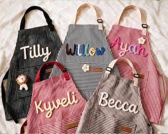 May include: Five aprons with vertical stripes in black, red, and brown. Each apron features a front pocket, an adjustable neck strap, and a leather-like tag labeled "apron." Names like Tilly, Willow, Ayah, Kyveli, and Becca are in raised lettering.