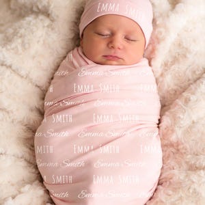 K&ouml;nnte beinhalten: Eine hellrosa Baby-Wickeldecke und eine passende M&uuml;tze, beide mit dem Namen Emma Smith in wiederholter wei&szlig;er Schrift bedruckt. Die Wickeldecke ist fest um einen schlafenden S&auml;ugling gewickelt und zeigt den weichen Stoff.