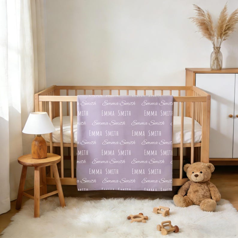 K&ouml;nnte beinhalten: Eine hellviolette Babydecke mit einem sich wiederholenden Muster des Namens Emma Smith in wei&szlig;em Druck. Die Decke h&auml;ngt &uuml;ber der Seite eines h&ouml;lzernen Kinderbetts und zeigt eine Mischung aus Schreibschrift und Blockschrift.