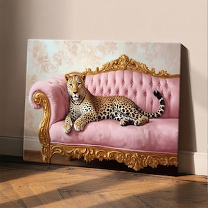 May include: A canvas print featuring a leopard resting on a pink tufted sofa with ornate gold trim. The background has a floral pattern. The leopard has a spotted coat and is looking directly at the viewer. The artwork is displayed in a room with wooden flooring.